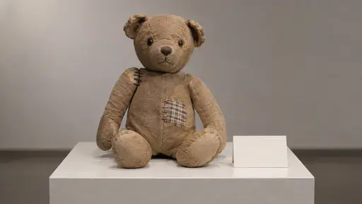 A worn cloth teddy bear sits on a museum plinth with one arm reattached by visible stitches and a checked cloth patch on its belly, a blank label card beside it.