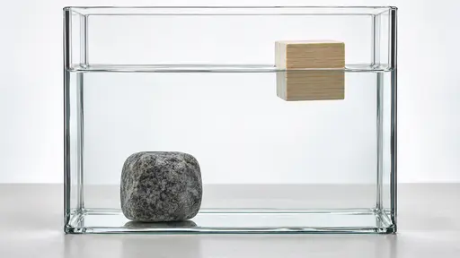 A grey granite stone lies on the floor of a wide clear glass tank of water on the left while a pale wooden block floats at the waterline on the right, in front of a bright diffused backdrop.