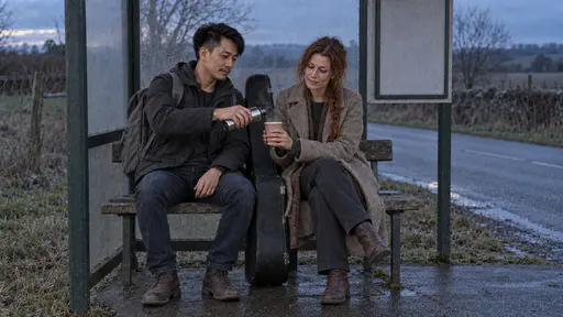 A wiry man in a charcoal jacket holds out a small steel flask to pour a steaming paper cup of tea for a freckled woman with a loose auburn braid beside him on a rural bus stop bench at dusk with her black guitar case leaning between them and a quiet country road behind.