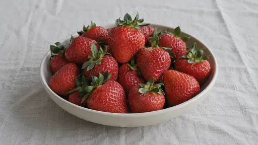 Whole strawberries with their green tops nestle together in one plain shallow dish on a plain cloth in bright even light.