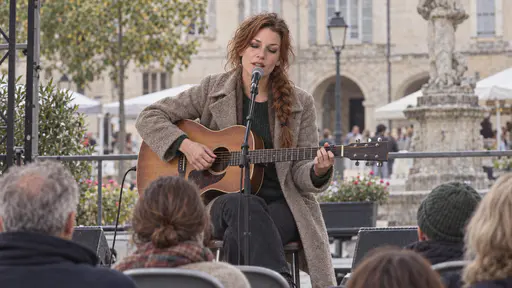 A freckled woman with a loose auburn braid and a silver nose ring in an oatmeal wool coat over a green jumper sits on a stool playing an acoustic guitar and singing with her eyes half closed on a small stage in a town square with a few rows of folding chairs and audience seen from behind.