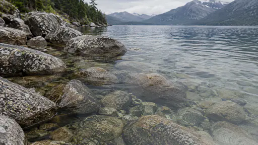 Rounded submerged boulders line the shore of a mountain lake with their forms visible through the transparent shallow water.