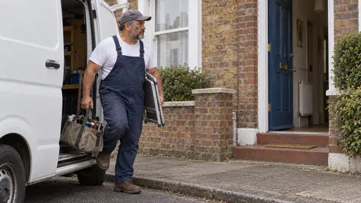 A stocky repair worker with a short grey beard in dark blue overalls and a grey cap steps down from a plain white van with a canvas toolbag in one hand and a folded step stool under his arm outside a small terraced house with a blue front door.
