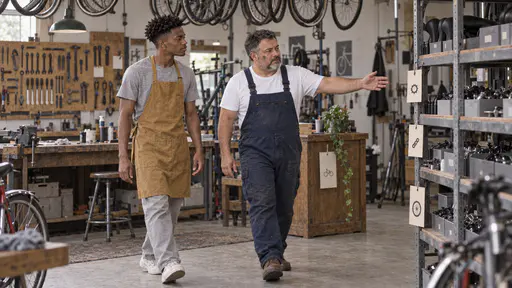 A stocky mechanic with a short grey beard in dark blue overalls points ahead with an open hand while a tall young man in a mustard apron follows with his eyes on the metal parts shelf in a small bicycle workshop in soft window light.