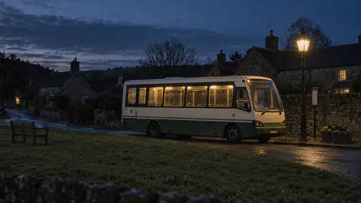 A small cream and green rural bus stands parked alone beside a village green at blue dusk with its interior lights glowing warm yellow and a street lamp lit beside it with dark cottages and a stone wall behind.