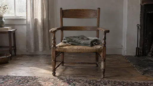 A worn oak armchair with a rush seat stands empty in a bare living room with a dented cushion, a folded blanket and a pair of reading glasses on the seat beside a half-drawn curtain and dust motes in overcast light.