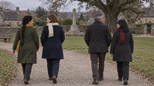 Four people in dark coats and coloured scarves walk away in twos along a gravel path seen from behind with a stone memorial and laid flowers small and warm in the long low evening light behind them.