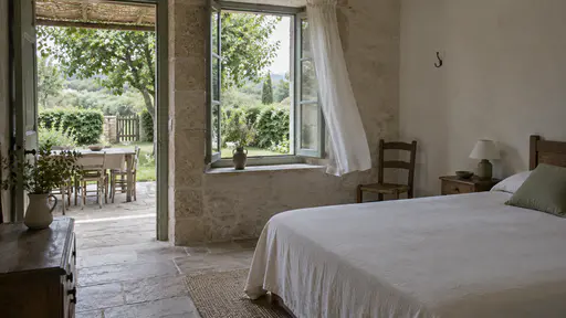 A cleared guest room with a bed made square, bare wall hooks, an empty chair and the window open to the green garden with the curtain lifting in pale morning light.