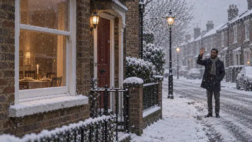 A tall bearded man in a navy duffel coat and grey scarf waves from a lamplit corner of a snowy street as snow falls while the lit window of a brick house in the foreground shows a laid table with a candle.