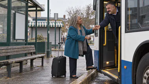 A heavyset moustached driver in a navy jacket leans out of an open bus door offering a hand to a woman with silver locs in a teal coat who stands on the kerb with her suitcase at her feet with a shuttered kiosk behind them in warm light.