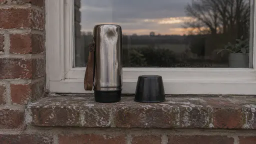 A dented steel flask stands upside down and empty beside an upturned black cup drying on a weathered brick windowsill with a field and a plant pot seen through the window in soft dusk light.