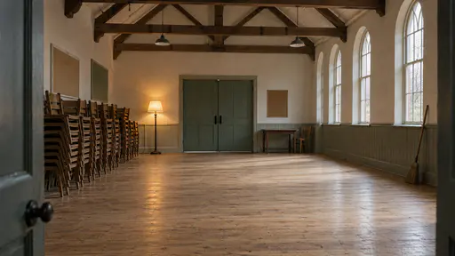 An empty timber-beamed community hall has wooden chairs stacked in neat columns along the wall, a swept pale floor, a broom leaning on the wall and one standing lamp lit in low warm light.