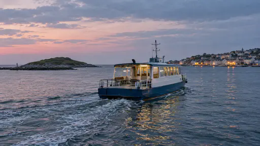 A small blue and white ferry with glowing warm yellow cabin windows crosses a calm strait with a pale wake at dusk between the low shape of a green island and the lights of a mainland harbour town.