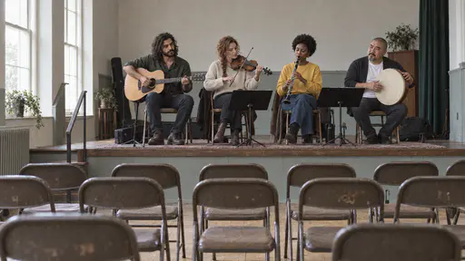 Four musicians with a guitar, a fiddle, a clarinet and a frame drum perform on a low stage in a community hall in bright side light from high windows with rows of empty folding chairs facing them.