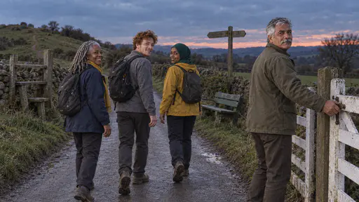 An older man in an olive jacket closes a white wooden field gate behind three walkers heading away down a lane glancing back at deep blue dusk with the field dark behind.