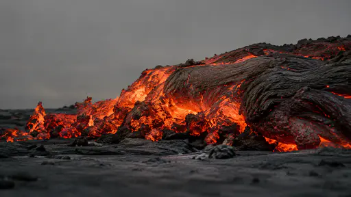The glowing front of an advancing lava flow pushes slowly over black ground in flat grey light, its leading edge crumbling into bright fragments.