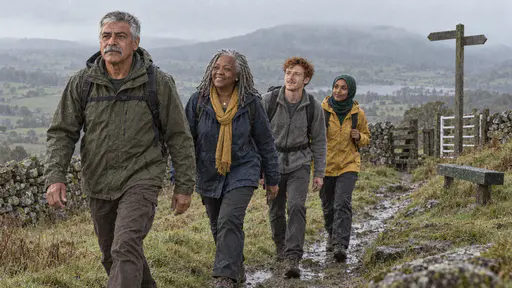 Four walkers in single file cross a wet grassy ridge path in light rain with a dry-stone wall and a far hill behind them under cool overcast light.