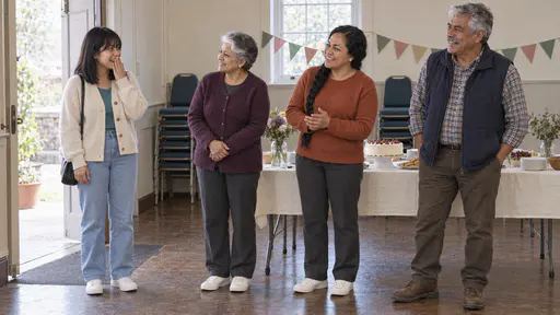 A young woman in a cream cardigan over a teal top stands just inside a hall doorway with a hand to her mouth in surprise while an older woman in a plum cardigan, a woman in a rust-orange jumper and an older man in a navy gilet turn smiling toward her beside a laid table.