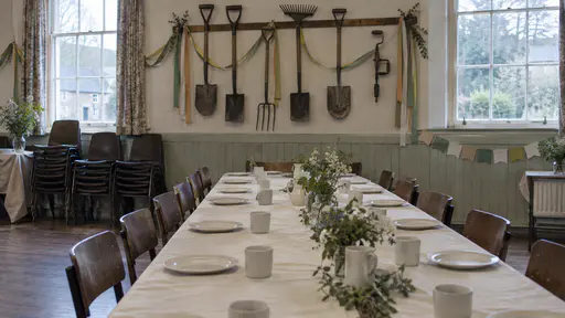 A village hall stands ready with a long table laid with a plain cream cloth, plates and cups and garden spades, rakes and a hand drill hung along the wall above as decoration with paper ribbons in cool morning light.