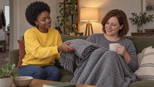 A young woman with a short natural afro in a bright yellow sweatshirt hands a folded grey blanket to a white woman with a chin-length auburn bob in a heather-grey sweater who sits curled on a sofa with a mug of tea and tucks it over her knees in warm lamp light in a tidy living room.