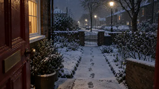 A single line of footprints leads from a dark red front door and glowing porch lantern down a cleared path through fresh snow toward a garden gate and dark street in cool blue night light.