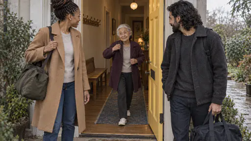 A very small elderly woman in a plum cardigan pulls on a coat and steps onto the threshold of an open yellow front door with a small smile as a tall woman and a bearded man wait on the path outside with bags in golden light.