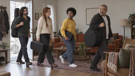 A bearded man with a guitar case, a woman with a red braid and a violin case, a young woman with a clarinet case and a bald man with a round drum bag enter a small living room with a rust-orange sofa in soft morning light.