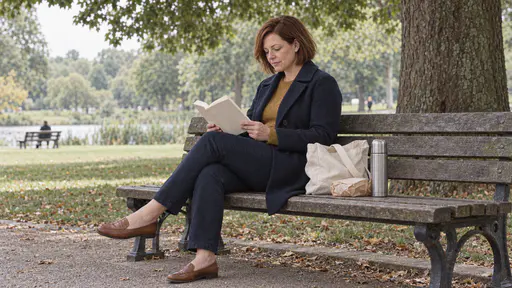 A woman with a chin-length auburn bob in a navy coat reads an open paperback on a park bench with an untouched wrapped sandwich and a thermos beside her in midday tree shade.