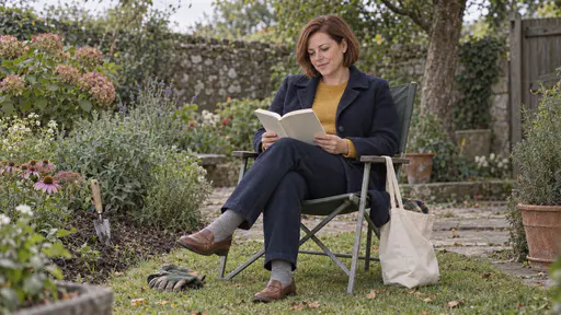 A woman with a chin-length auburn bob in a navy coat reads an open paperback in a folding garden chair with a pair of gardening gloves dropped at the chair leg and a trowel in the flowerbed beside her in dappled garden light.