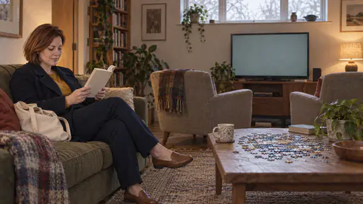 A woman with a chin-length auburn bob in a mustard jumper reads a paperback at one end of a sofa while two empty armchairs face a muted blank television across a living room with a half-finished jigsaw on the coffee table in warm lamp light.