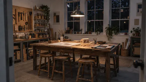 A long wooden worktable in an empty community room at night holds a half-finished sketch, a small wooden model, a folded patchwork square and a curl of wood shavings under a hanging lamp left glowing with a stool pushed in at each place.