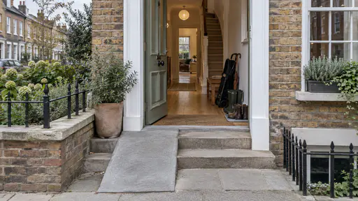 A sage green front door stands open beside a gentle stone ramp and two steps with a folded pushchair, a walking stick, a bench and a tray of boots of different sizes just inside in soft overcast light.