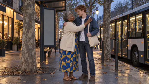 A young man in a denim jacket hugs an older woman in a cobalt dress goodbye at a bus-stop corner under a plane tree at blue dusk with a lit bus waiting behind them and shop windows glowing warm.