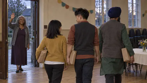 A young woman in a mustard cardigan, a man in a rust shirt and a man in a navy turban walk away down hall steps from behind each carrying a plain brown-paper parcel while a woman in a purple dress waves from the lit doorway in low evening light.