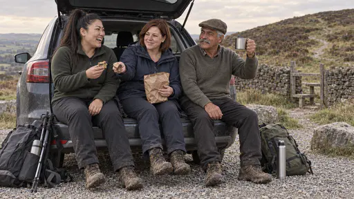 Three hikers, a woman with an auburn bob in a navy jacket, a broad-shouldered woman with a high ponytail in a green fleece and an elderly man in a flat cap, sit on an open car boot with muddy boots passing cake and raising a tin cup in a toast at a gravel car park.