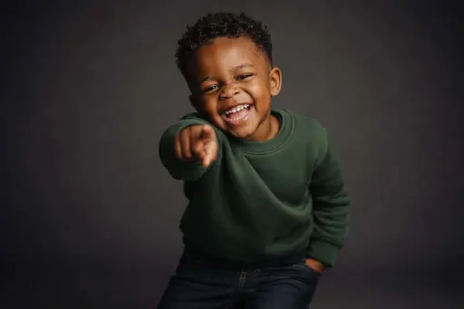 Cinematic 3:2 editorial studio portrait of a grinning three-year-old Black boy with tight curls leaning forward and pointing at the lens in a forest-green sweater on a seamless deep-charcoal backdrop with warm amber highlights.