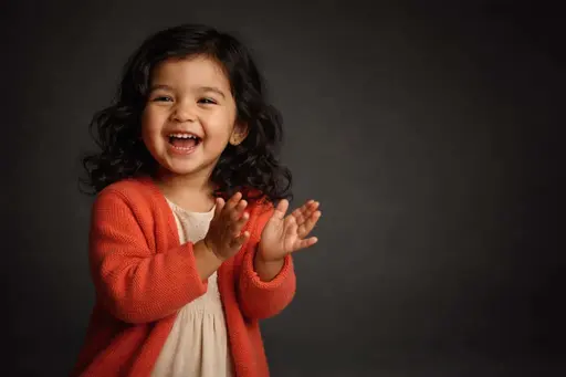 Cinematic 3:2 editorial studio portrait of a laughing three-year-old Latina girl in a coral cardigan and cream dress on a deep charcoal seamless backdrop, hands mid-clap with soft wrapped light, subtle rim, shallow depth and gentle film grain.