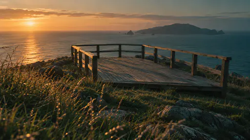 An empty timber lookout platform with a low railing stands on a grassy headland above a calm sea and a distant island in low warm sunset light.