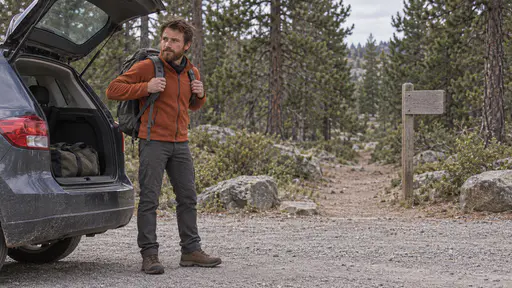 A lean man with an auburn beard in a rust fleece and walking boots settles a grey rucksack onto his shoulders beside the open boot of a small hatchback at a trailhead car park at the edge of a pine forest with a trail opening between the pines and a blank marker post in soft afternoon light.