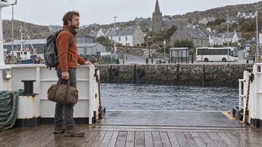 A lean man with an auburn beard in a rust fleece with a grey rucksack stands at the open gangway of a small ferry at a quay holding a small brown duffel bag in one hand and the rail in the other looking toward a harbour town and a bus waiting at the stop beyond in soft grey light.