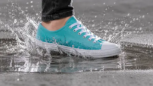 A foot in a turquoise canvas low-top sneaker with white laces and a white rubber toe cap, below a dark jeans cuff, stepping into a shallow puddle on grey concrete, a thin crown of water and droplets frozen around the sole and ripples spreading.