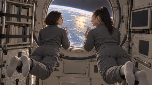 A slim woman with a sleek dark bob and a powerfully built woman with a thick black high ponytail, both in plain slate-grey crew jumpsuits, float side by side holding warm drink pouches with straws looking out of a large round habitat window at a bright blue planet below in soft evening-coloured light.