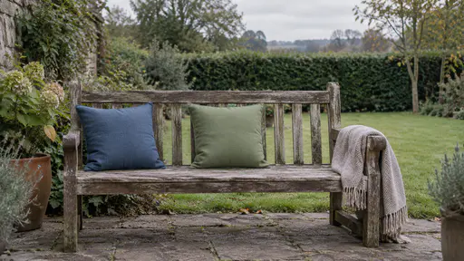 A wooden garden bench has a blue cushion pushed neatly to one end and a green cushion placed beside it with a clear gap between them and a folded blanket on the arm with a lawn and hedge behind in soft morning light.