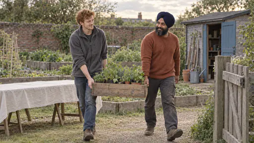 A young man with copper-red hair in a grey fleece and a broad man in a navy turban and rust-orange jumper carry a wooden crate of potted seedlings between them toward a garden gate in late golden light.