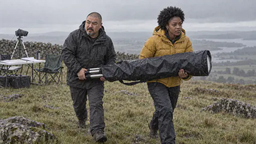 A stocky shaven-headed man in a charcoal parka and a young woman in a mustard puffer jacket carry a white telescope wrapped in a waterproof cover between them across a rainy hilltop field with rain streaking diagonally.