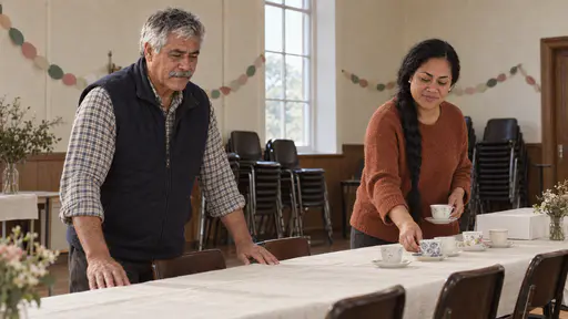 An older man in a navy gilet smooths a plain cream cloth along a long trestle table while a woman with a long braid in a rust-orange jumper sets out mismatched teacups and a plain white cake box in a community hall in warm light.