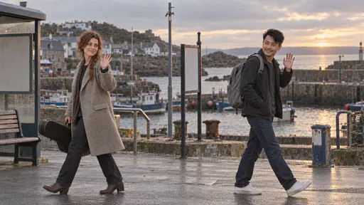 A freckled woman with a loose auburn braid carrying a black guitar case lifts a hand in a small wave over her shoulder as she walks away and a wiry man in a charcoal jacket raises a hand in a wave back as he shoulders a grey rucksack toward the other way with a harbour glinting behind them in golden evening light.