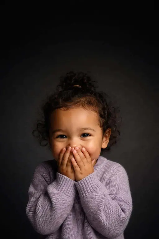 Cinematic 2:3 editorial studio portrait of a giggling two-year-old mixed-race girl with soft curls and medium tan skin, hands partly covering her mouth, lavender sweater, and a near-black gradient backdrop.
