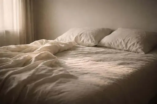 Quiet editorial 3:2 interior of an unmade bed bathed in warm, muted window light with rumpled sheets, film grain, soft haze, restrained palette, and generous negative space for icons.