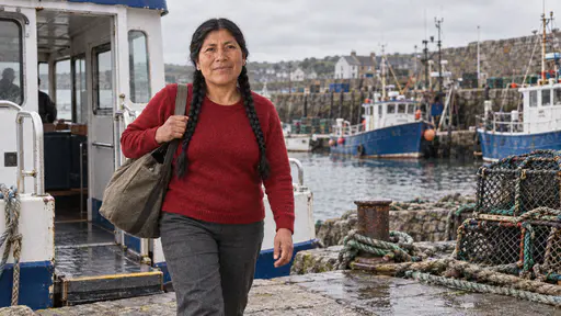 A sturdy woman with two long black braids in a deep red wool sweater steps off a small ferry onto a stone harbour quay with a canvas holdall on her shoulder, coiled ropes, lobster pots and blue and white fishing boats behind her in bright morning light.